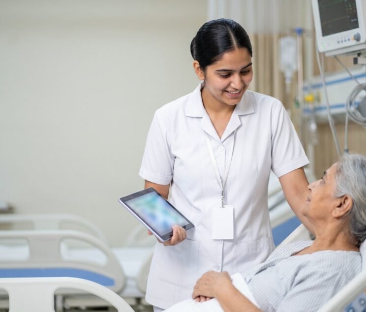 Nurse using a tablet at a patient's bedside while speaking kindly with the patient in an Indian hospital ward