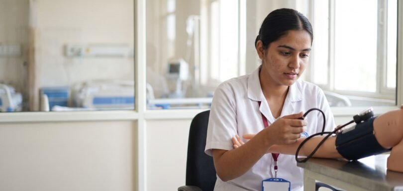 Nursing student practising blood pressure on a training mannequin in a skills lab with a hospital ward behind her